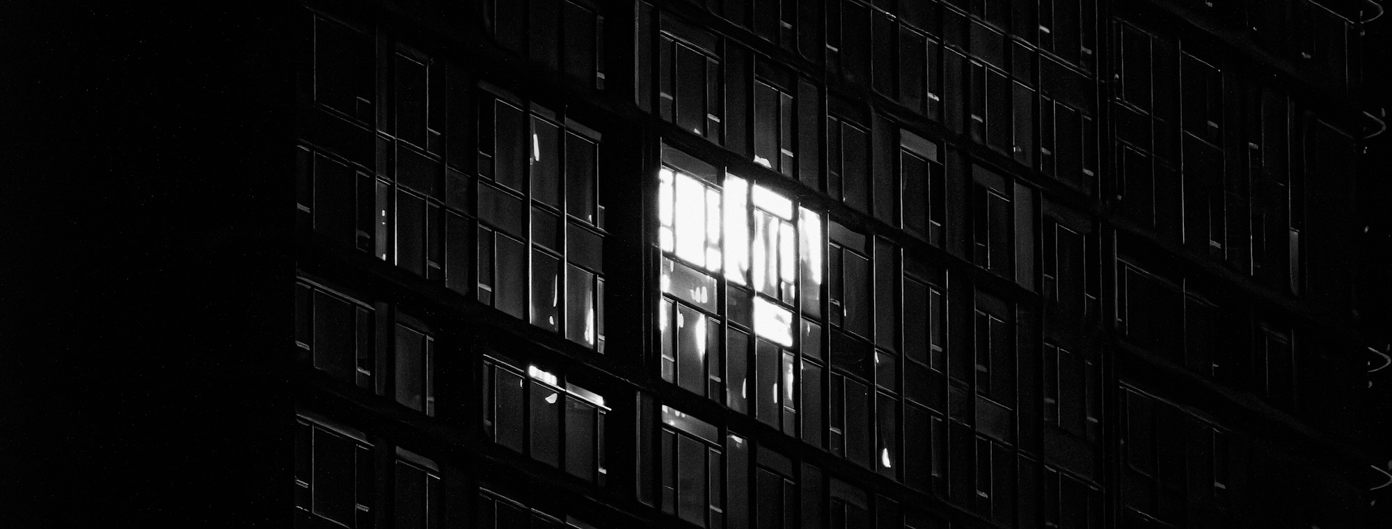 A tower at night. Almost every window is dark and one of them is lit.